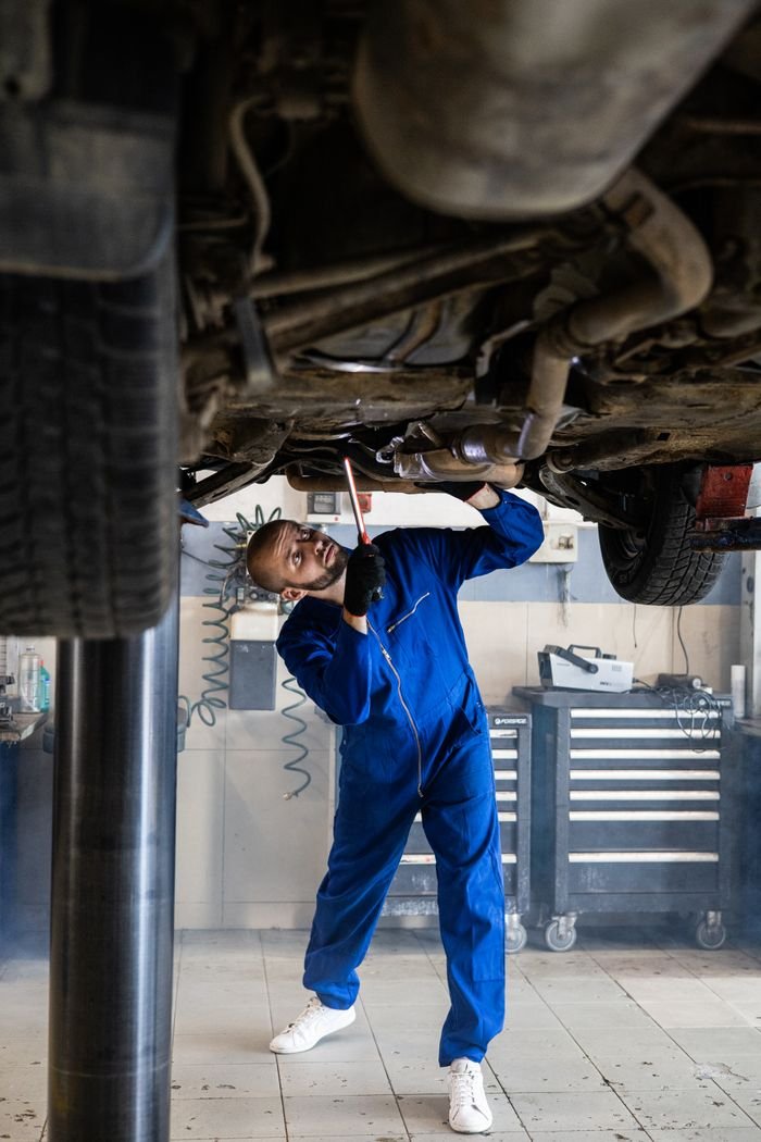 A female mechanic in blue coveralls inspects a cars undercarriage in an auto repair shop.